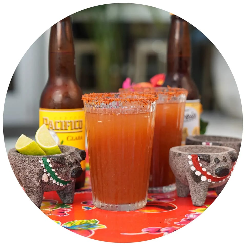 Two micheladas with chili-lime rims, lime wedges in a molcajete, and Pacifico beer bottles.