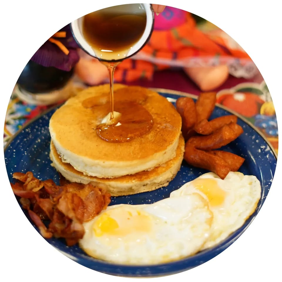 Maple syrup being poured over a stack of pancakes, with bacon, sausage, and fried eggs.