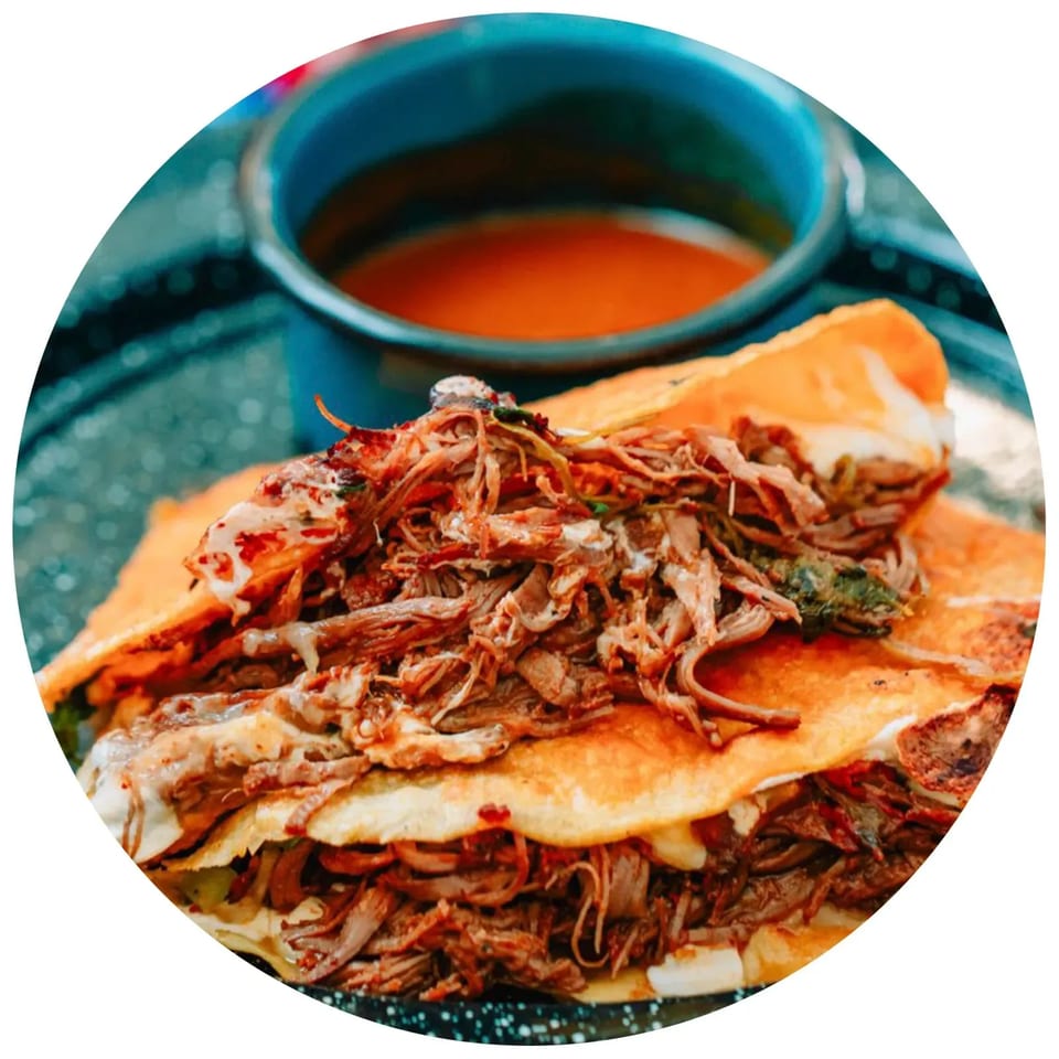 Close-up of two birria tacos overflowing with shredded meat and cheese, with a side of consommé.