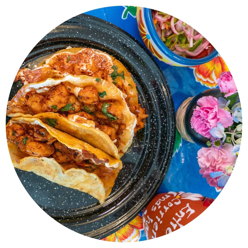 Three shrimp tacos with crema and cilantro on a black plate, with a side of pickled onions and flowers.